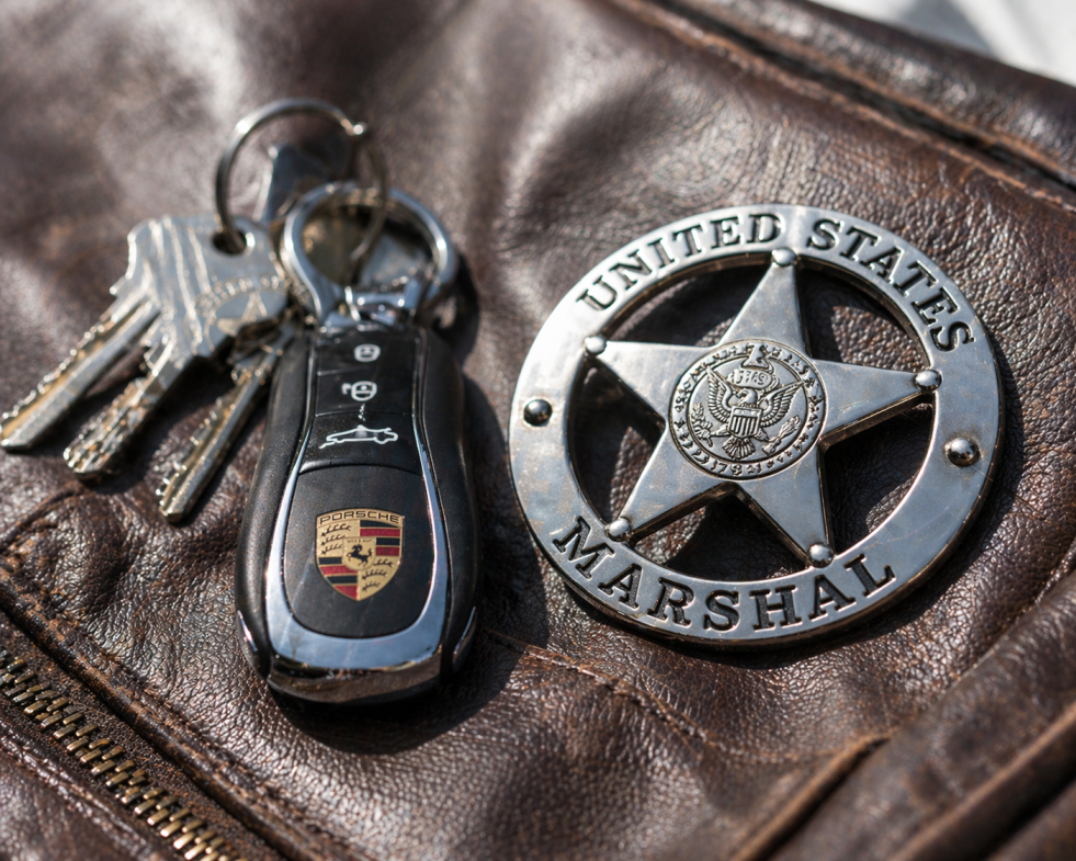 A close-up of a U.S. Marshal badge and Porsche keys on a leather jacket, referencing Lucas Davenport from John Sandford's Revenge Prey.